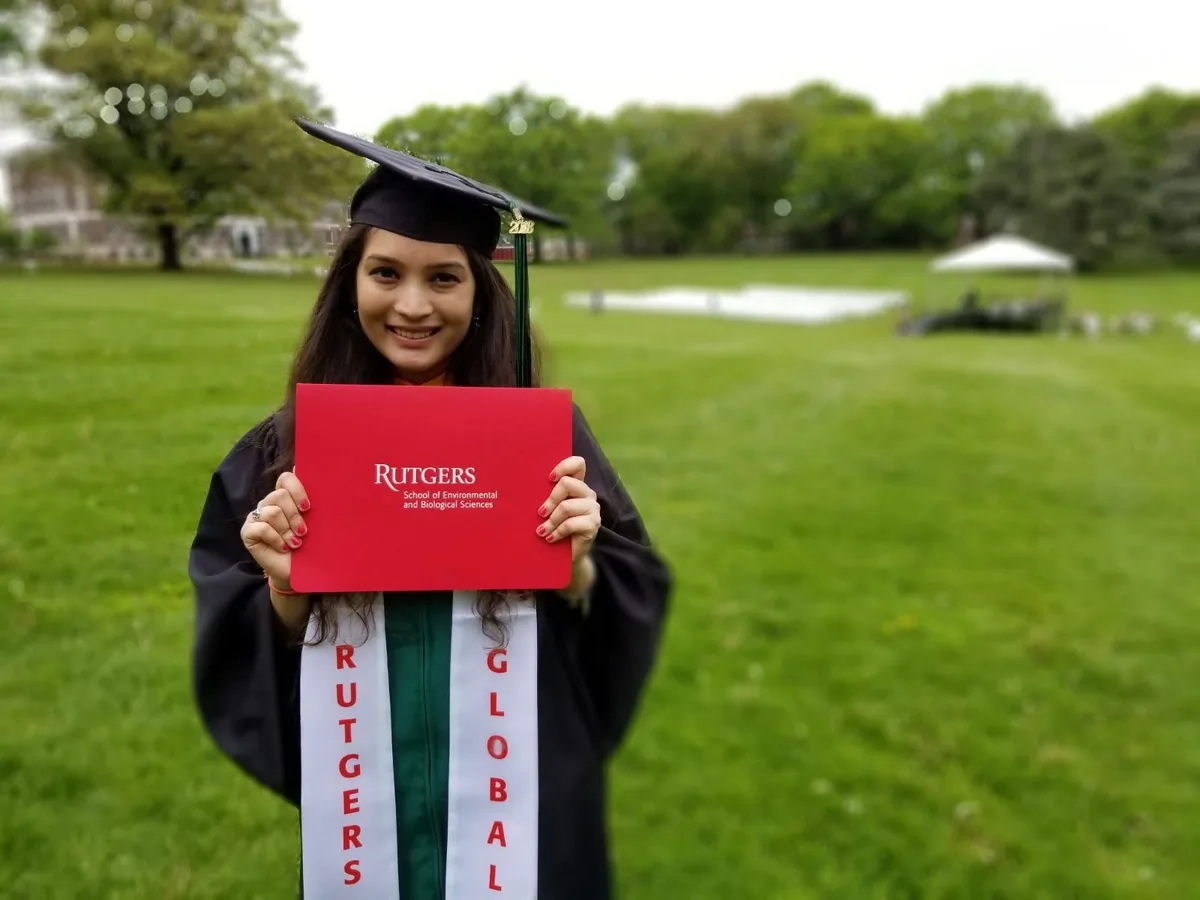 Riddhi Jain in a graduation gown holding a Rutgers diploma cover on a lawn